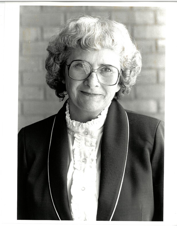 Dr. Patricia McIlrath, an older woman, poses for a photograph. She has white hair and wears glasses, a jacket, and a shirt with ruffles.