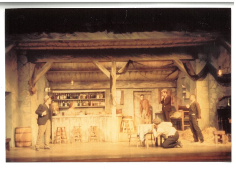 Five men and a woman perform a scene from The Playboy of the Western World on a set resembling a rustic bar. One of the men crouches behind a table. The woman and one of the men stand on the left side of the stage, facing the other three men on the right side of the stage.
