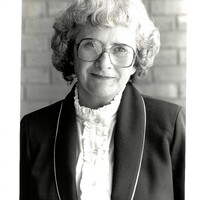 Dr. Patricia McIlrath, an older woman, poses for a photograph. She has white hair and wears glasses, a jacket, and a shirt with ruffles.