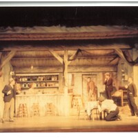 Five men and a woman perform a scene from The Playboy of the Western World on a set resembling a rustic bar. One of the men crouches behind a table. The woman and one of the men stand on the left side of the stage, facing the other three men on the right side of the stage.